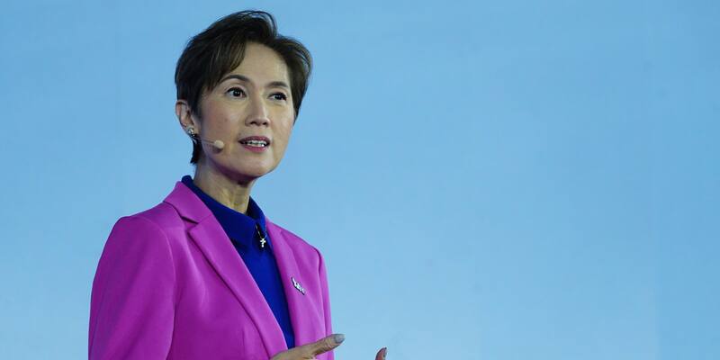 Josephine Teo in a magenta jacket and blue blouse speaking on stage against a pale blue background