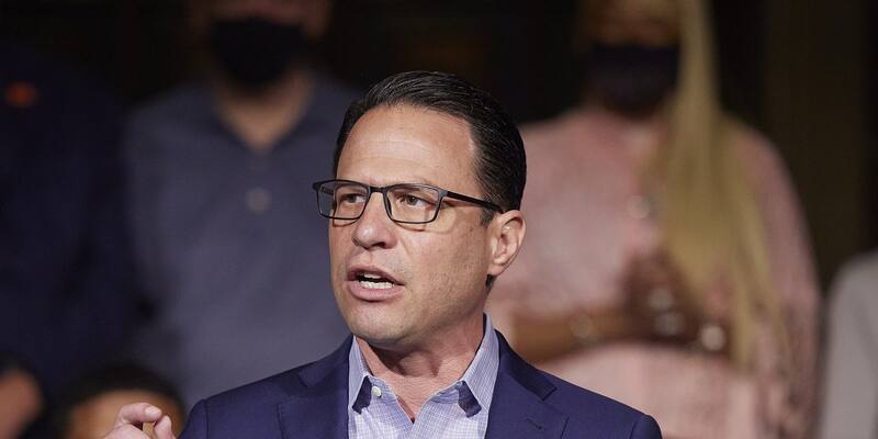 Josh Shapiro in glasses, a navy jacket and a checked shirt speaks at a microphone, with people in face masks blurred behind him