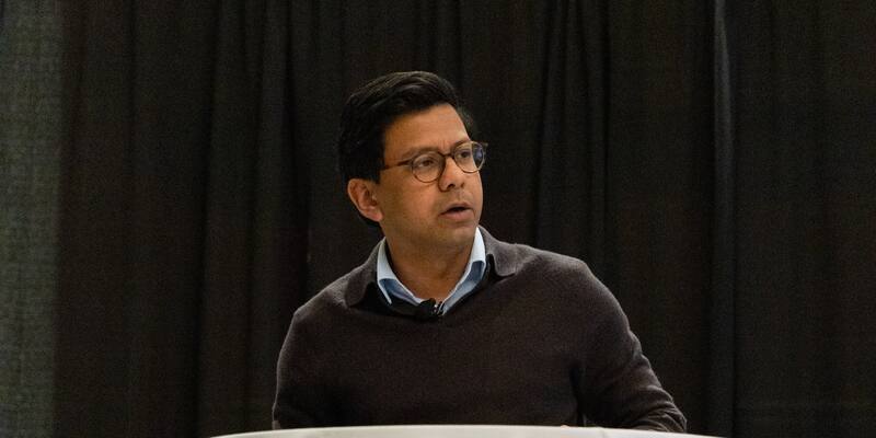 Kanishka Narayan, wearing glasses, a dark jumper and a light blue shirt, speaking at a lectern in front of a black curtain