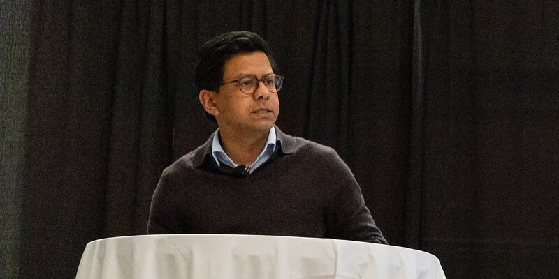 Kanishka Narayan, wearing glasses, a dark jumper and a light blue shirt, speaking at a lectern draped in a white cloth in front of a black curtain
