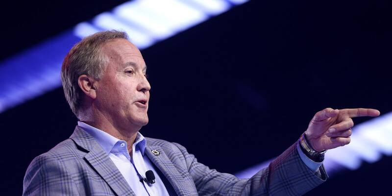 Ken Paxton, in a grey checked jacket and open-necked shirt, points to his left while speaking on a stage lit in blue