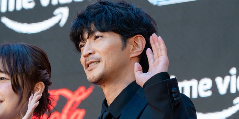 Voice actor Kenjiro Tsuda, in a black suit and shirt, smiling and waving on a red carpet in front of a large screen