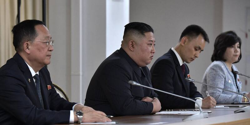 Kim Jong Un in a dark suit sits at a conference table between North Korean officials, with microphones in front of them