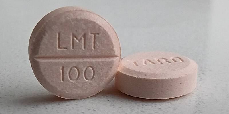 Two pale pink lamotrigine tablets, one standing upright marked LMT 100, on a white speckled surface