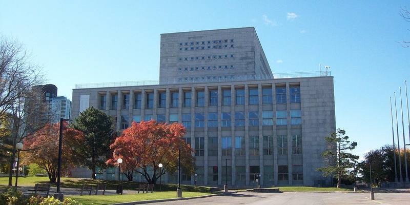 A large grey stone building with rows of tall windows and a taller block behind it, with autumn trees and lamp posts in front under a blue sky