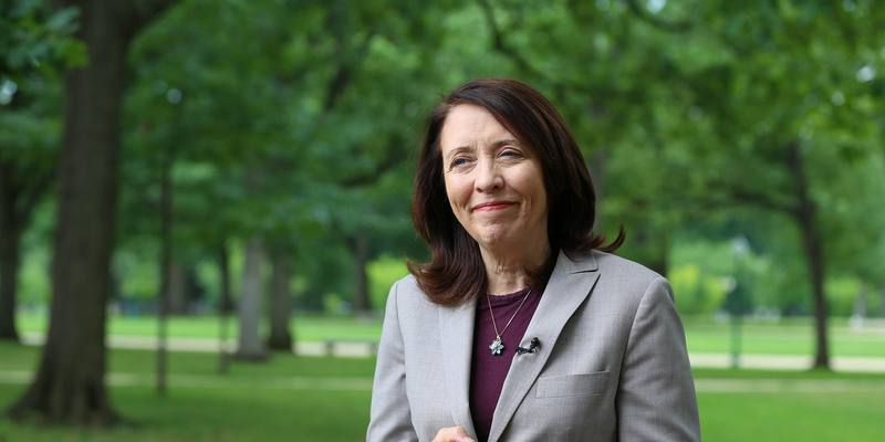 Senator Maria Cantwell, in a grey jacket and burgundy top, smiling outdoors in a park with green trees behind her