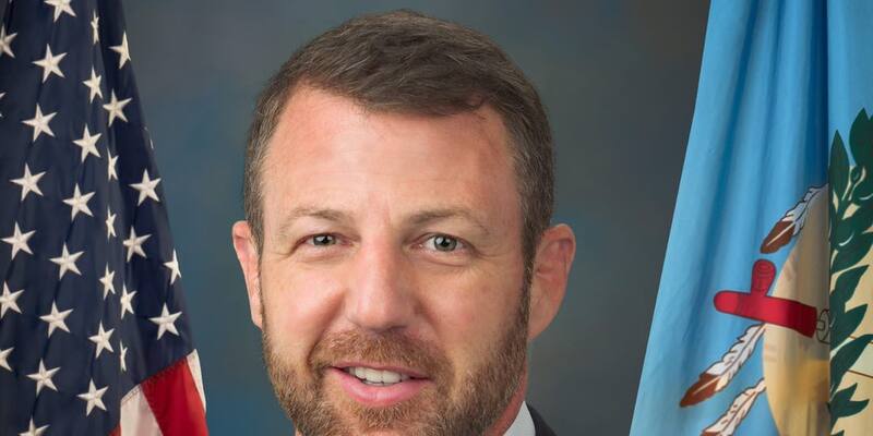 Markwayne Mullin in a dark suit and red striped tie, smiling, in front of the US and Oklahoma flags