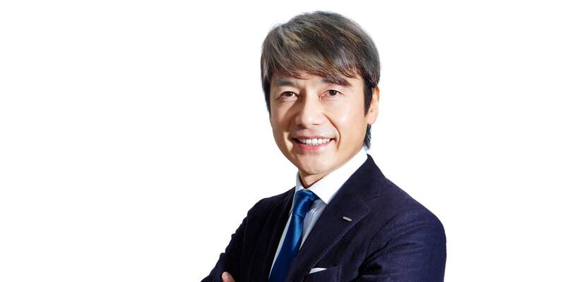 Masatoshi Kumagai, smiling, in a dark navy suit and blue tie with arms folded, against a white background