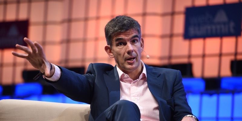 Matt Brittin in a navy jacket and pink shirt, gesturing with one hand as he speaks on stage, with a Web Summit sign behind him