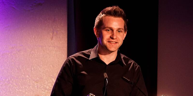 Max Schrems in a black shirt smiling as he speaks at a lectern with microphones on a purple-lit stage