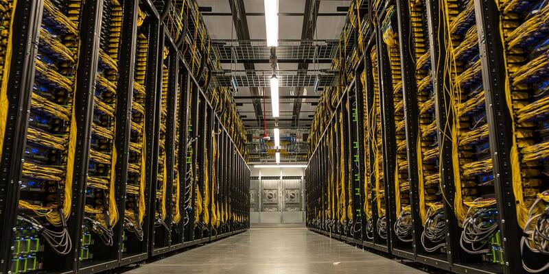 Two long rows of black server racks threaded with yellow fibre cables either side of a lit aisle inside a Meta data centre