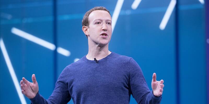 Mark Zuckerberg in a blue jumper speaking on stage with his hands spread against a blue backdrop