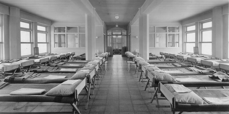 A black and white archive photo of rows of empty, neatly made hospital beds either side of a tiled central aisle.