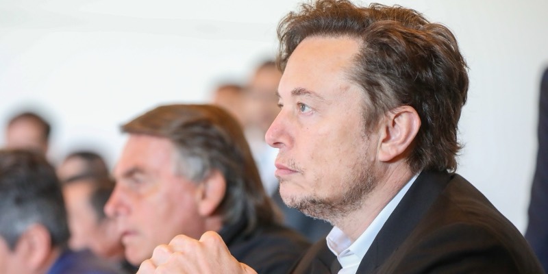Elon Musk in profile in a dark jacket, hands clasped, listening intently, with other attendees blurred behind him