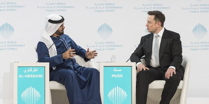 Elon Musk in a dark suit and grey tie sits in a white armchair facing Mohammad Al Gergawi, in white headdress and blue robe, on a World Government Summit stage behind name plates reading Al-Gergawi and Musk