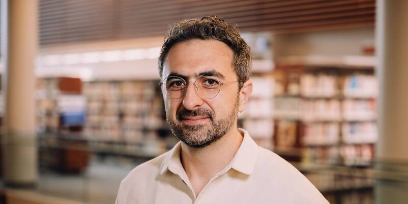 Mustafa Suleyman, in round glasses and a cream shirt, smiles in front of library shelves