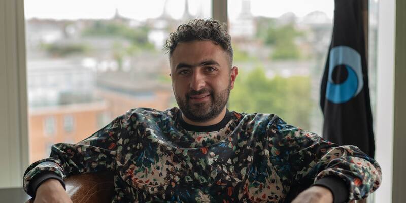 Mustafa Suleyman, in a patterned sweatshirt, sits in a leather chair by a window with a DeepMind flag behind him