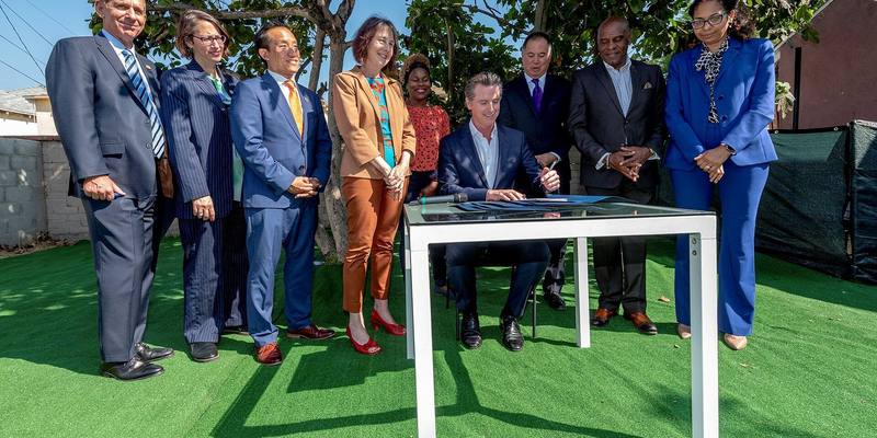 Gavin Newsom seated at a glass table signing documents outdoors on artificial grass, with eight lawmakers and officials standing behind him under a tree