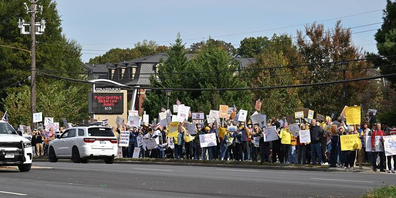Protesters holding handmade signs line a roadside in North Bethesda beside a Strathmore sign, with autumn trees behind