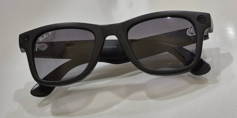 Black Ray-Ban Meta Wayfarer smart glasses on a white surface, with small camera lenses at both top corners of the frame
