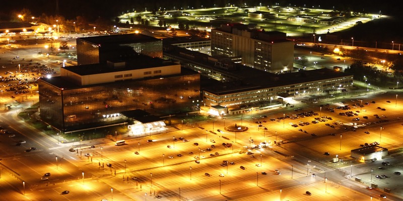 Aerial night view of the National Security Agency headquarters, with dark glass buildings surrounded by brightly lit car parks