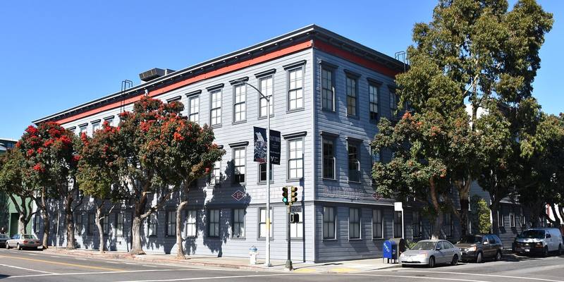 A three-storey grey wooden corner building with a red stripe under the roof and PIONEER BUILDING painted on the side, lined with flowering red trees under a blue sky