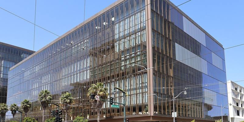 A glass-fronted office block with bronze vertical fins on a sunny street corner, with palm trees and traffic lights in front