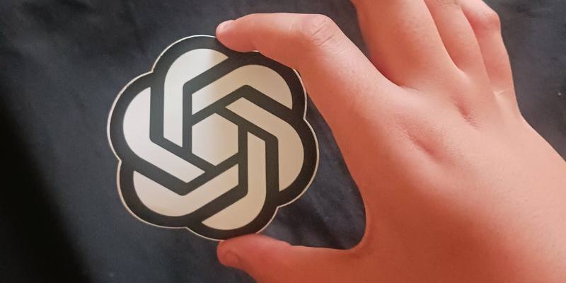 A hand holding a round black and white OpenAI logo sticker against a dark fabric background.