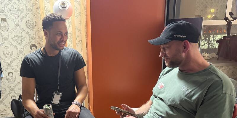 Peter Steinberger, in a black ClawCon cap and green T-shirt, looks at his phone while seated next to Tomas Taylor, who smiles in a black T-shirt and lanyard, against an orange wall