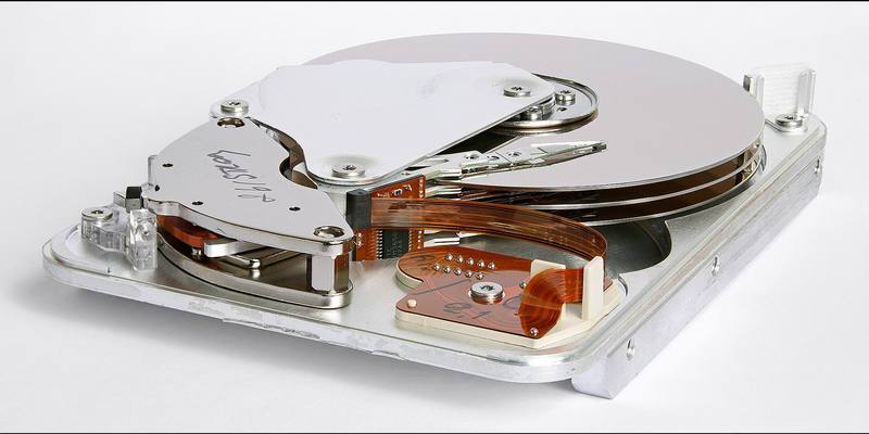 An opened hard disk drive on a white background, with shiny metal platters, the read/write arm and an orange ribbon cable.