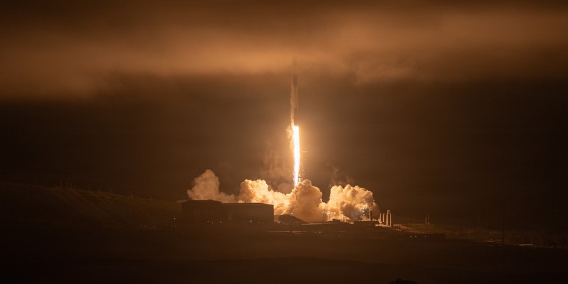 A Falcon 9 rocket lifts off at night from Vandenberg, its bright exhaust lighting clouds of smoke and a low orange sky