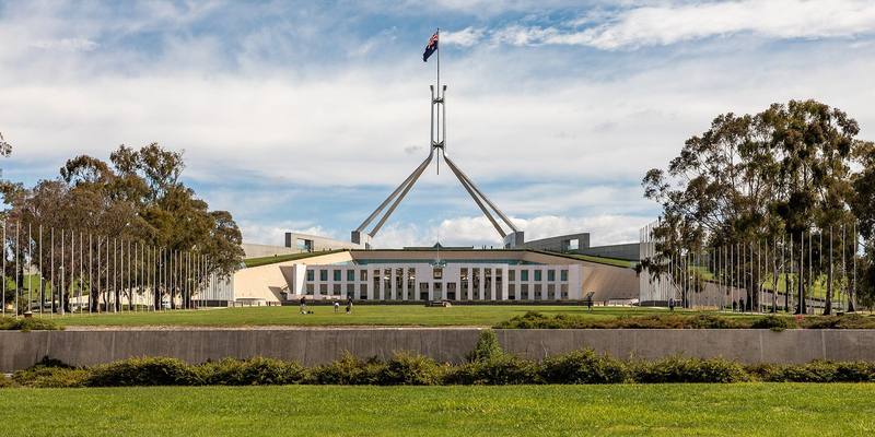 Australia's Parliament House in Canberra with its tall flagpole, lawns and eucalyptus trees under a cloudy blue sky