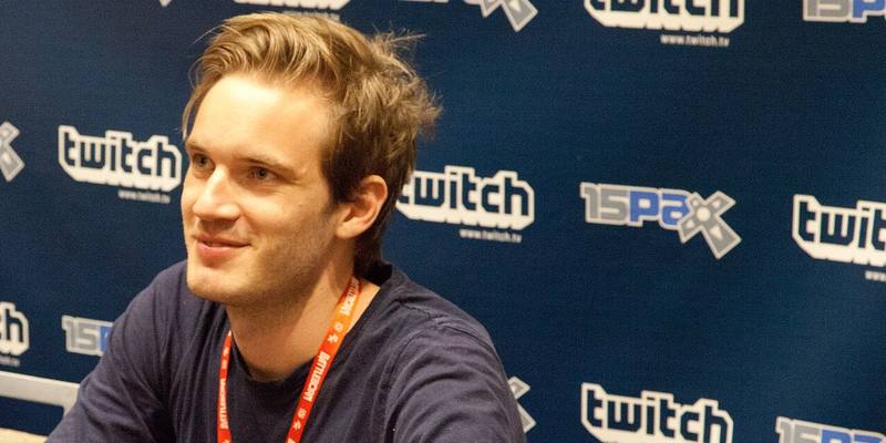 PewDiePie, smiling, in a navy top and red lanyard, seated in front of a Twitch and PAX backdrop.