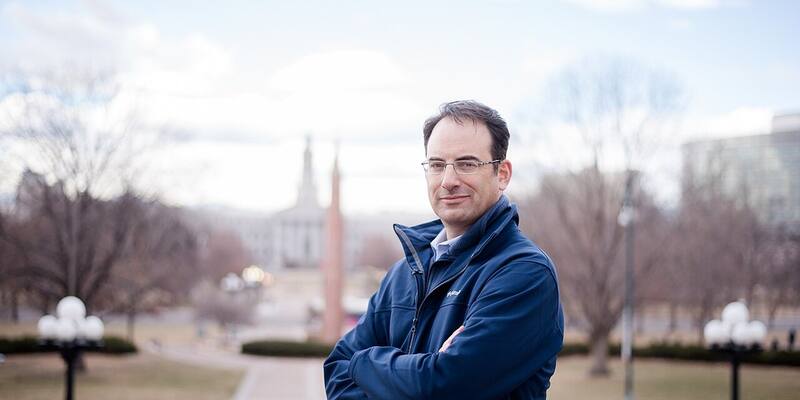 Phil Weiser in glasses and a navy zip-up jacket, arms folded, in a park with bare trees and Denver’s City and County Building blurred behind him