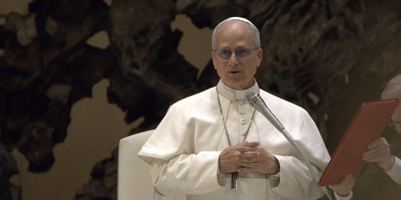 Pope Leo XIV in white robes and skullcap, hands clasped, speaking into a microphone