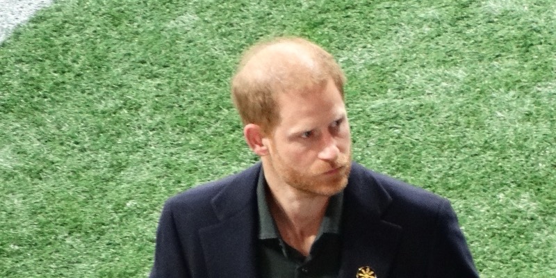 Prince Harry, in a dark coat, standing on a football field