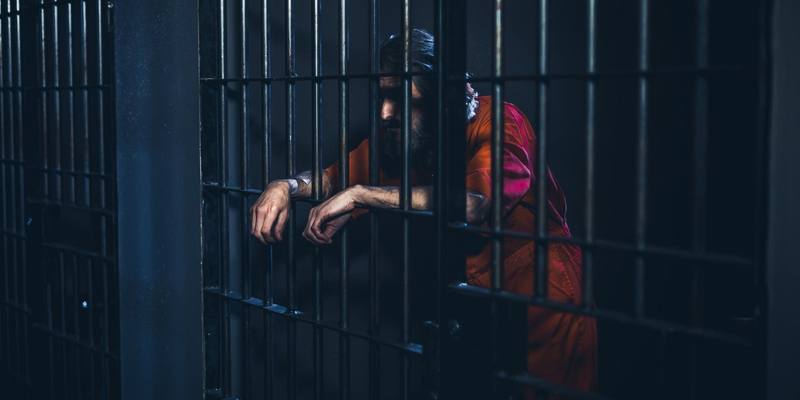 A bearded man in an orange prison jumpsuit leans his arms through the bars of a dark prison cell