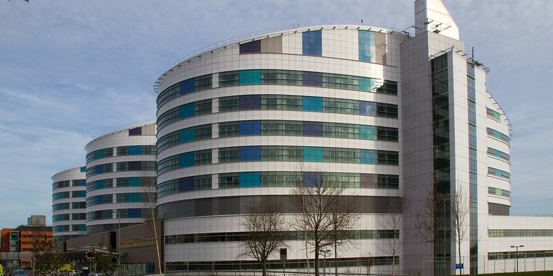 A large modern hospital building with curved, multi-storey wings of white panels and blue and green windows under a pale blue sky.