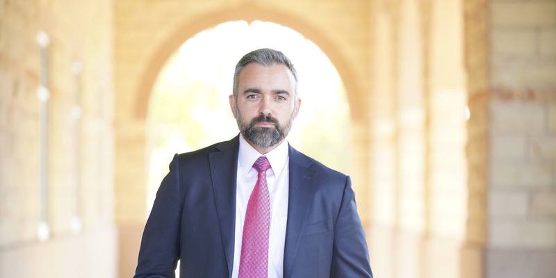 New Mexico Attorney General Raúl Torrez, in a navy suit and pink tie, standing in a sunlit stone arcade.