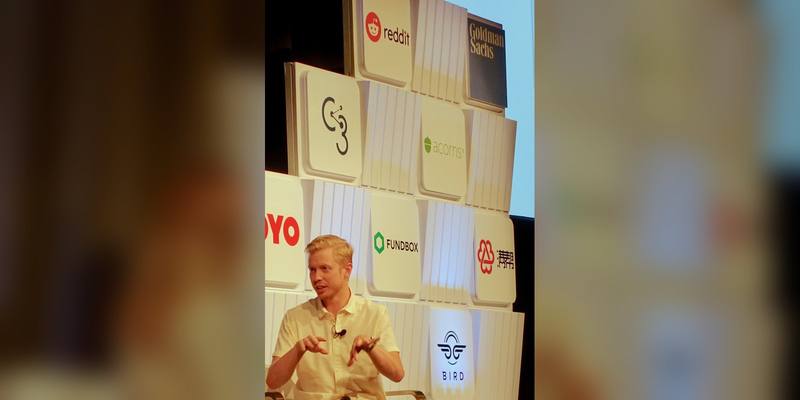 Reddit CEO Steve Huffman, in a cream short-sleeved shirt, gestures as he speaks on stage in front of a wall of company logos including Reddit’s