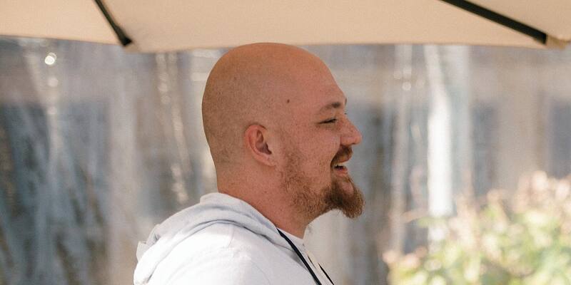 Amjad Masad, bald with a red beard and a white hoodie, laughs under a cream parasol at an outdoor event