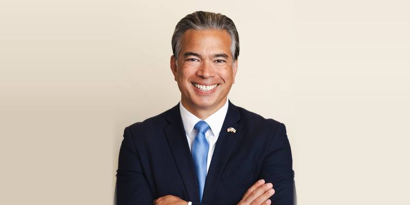 Rob Bonta, smiling with his arms folded, in a dark suit and light blue tie against a cream background
