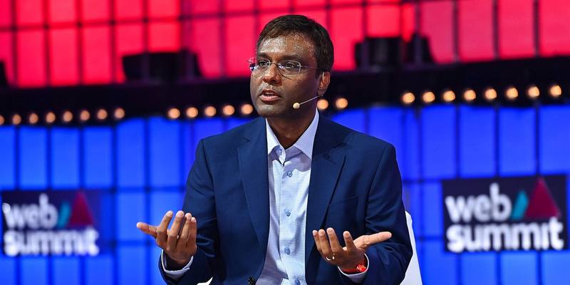 Rohit Prasad in a navy suit and glasses, gesturing with both hands as he speaks on stage in front of red and blue Web Summit screens.
