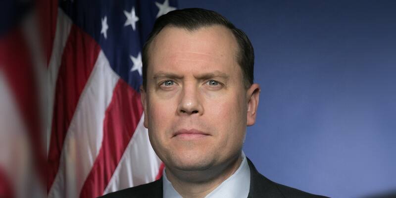 A man with short dark hair in a dark suit, white shirt and striped green and navy tie, in front of a US flag and a blue backdrop