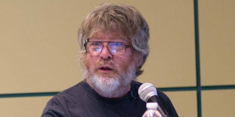 Russell Impagliazzo, a grey-haired, bearded man in glasses and a dark T-shirt, speaking into a microphone at a lectern