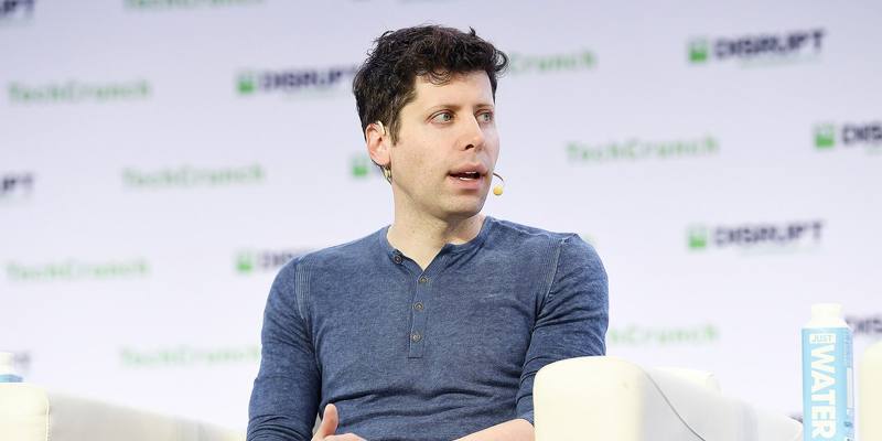 Sam Altman, in a blue long-sleeved henley and wearing a headset microphone, speaking from a white armchair in front of a TechCrunch Disrupt backdrop