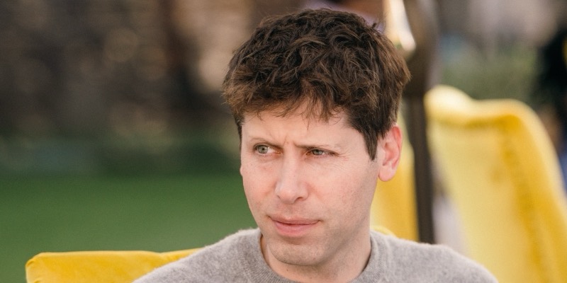 Sam Altman, frowning slightly and looking to one side, in a grey jumper on a yellow chair outdoors