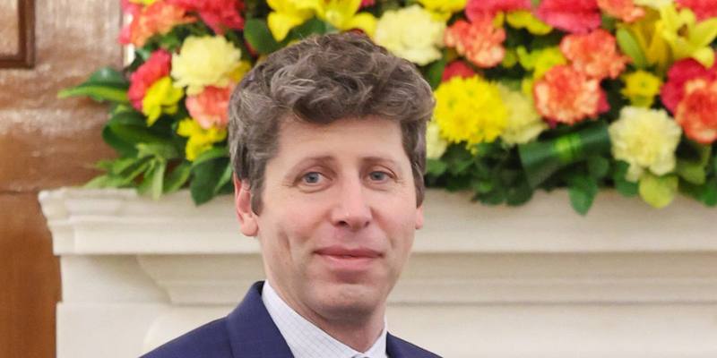 Sam Altman, in a navy suit, smiling in front of a mantelpiece covered in bright flowers