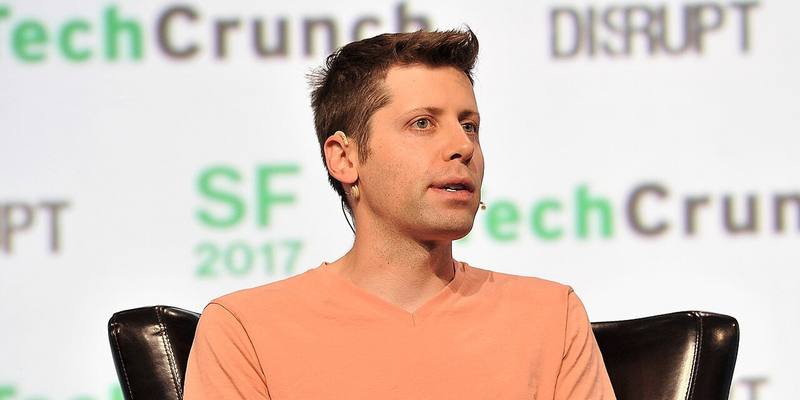 Sam Altman in an orange T-shirt, seated in a black leather chair and speaking, in front of a TechCrunch Disrupt SF 2017 backdrop.