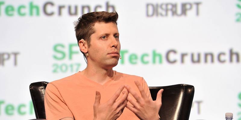 Sam Altman, in a salmon-coloured T-shirt, gesturing with both hands as he speaks from a black leather armchair in front of a TechCrunch Disrupt backdrop